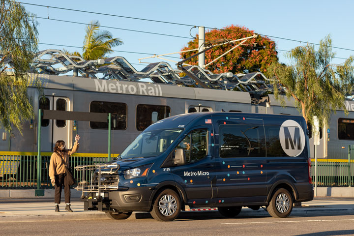 Metro Micro van in front of a Metro Rail train at a station with a person hailing the vehicle curbside.